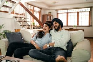 best movies for laughter, Happy young Indian man in turban and positive woman in casual clothes embracing and laughing while watching comedy movie sitting on sofa