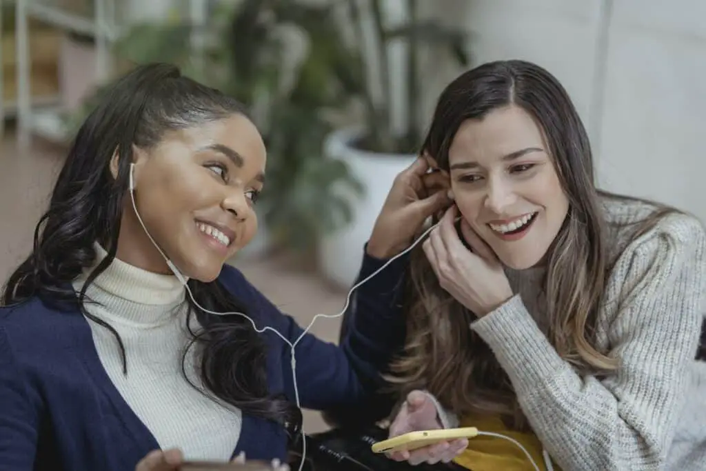 happy songs, Happy young African American lady with smartphone and earphones smiling and sharing music with female best friend while chilling together in cozy cafe